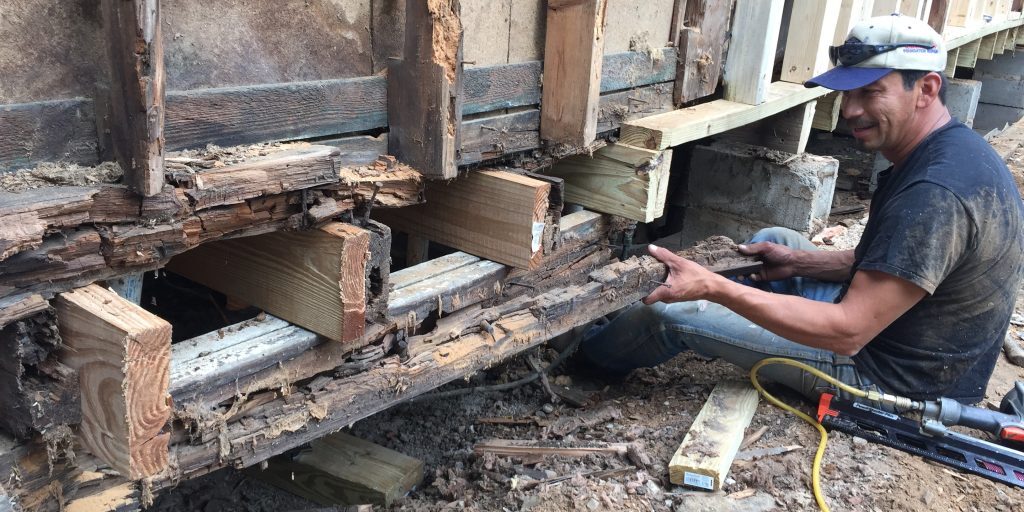 Contractor repairing rotted sill plate and rim joist, installing new pressure-treated lumber along a home’s exterior wall